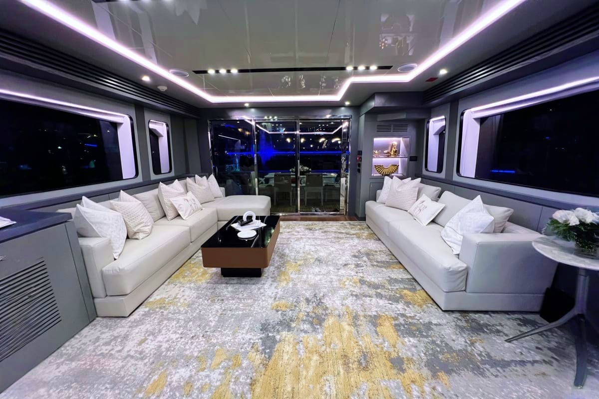 Luxury yacht interior with white sectional sofas, LED ceiling lighting, floor-to-floor windows, and ocean view at night