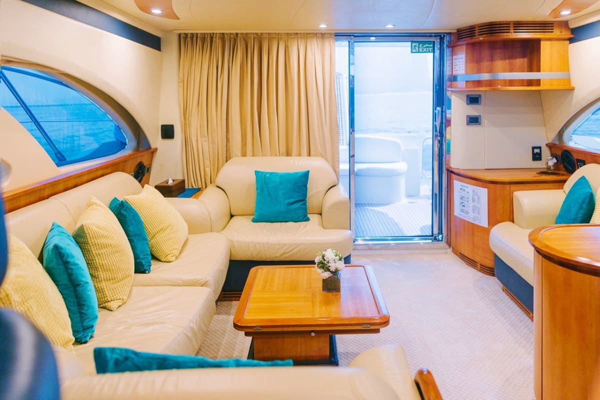 Luxury yacht interior with cream-colored seating, teal accent pillows, wooden furnishings, and ocean view through porthole wi
