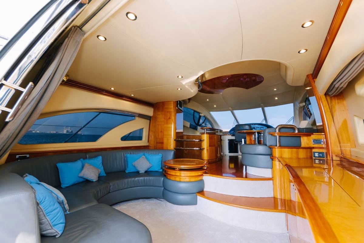Luxury yacht interior cabin with curved gray seating, blue pillows, wooden cabinetry, recessed lighting, and ocean views thro