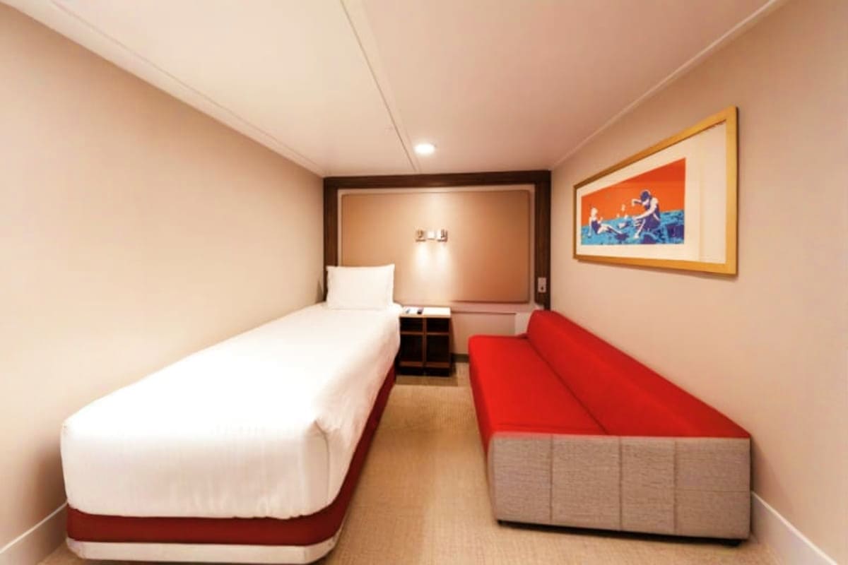 Genting Dream Singapore Roundtrip Cruise Spacious cruise ship cabin with single bed, red sofa, nightstand, and framed artwork on beige walls