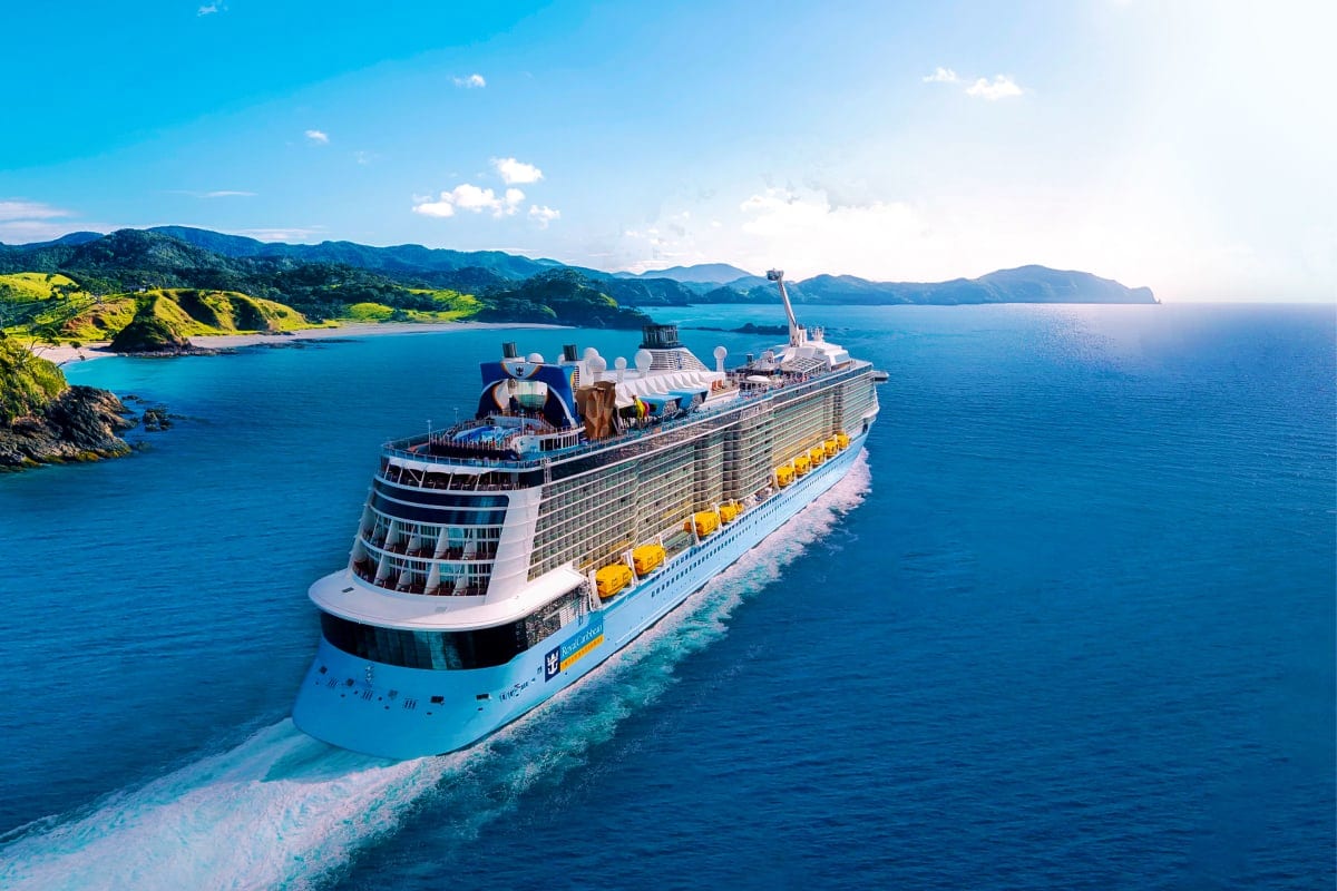 Large cruise ship with blue hull and yellow lifeboats sailing in turquoise tropical waters near green mountainous coastline u