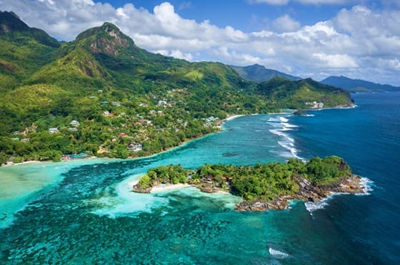 Aerial view of Seychelles island with turquoise lagoon, white sand beach, lush green mountains, and coastal village nestled a