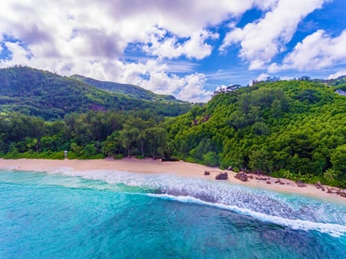 Aerial view of a pristine tropical beach with turquoise waters, white sand, and lush green forested hills in Seychelles