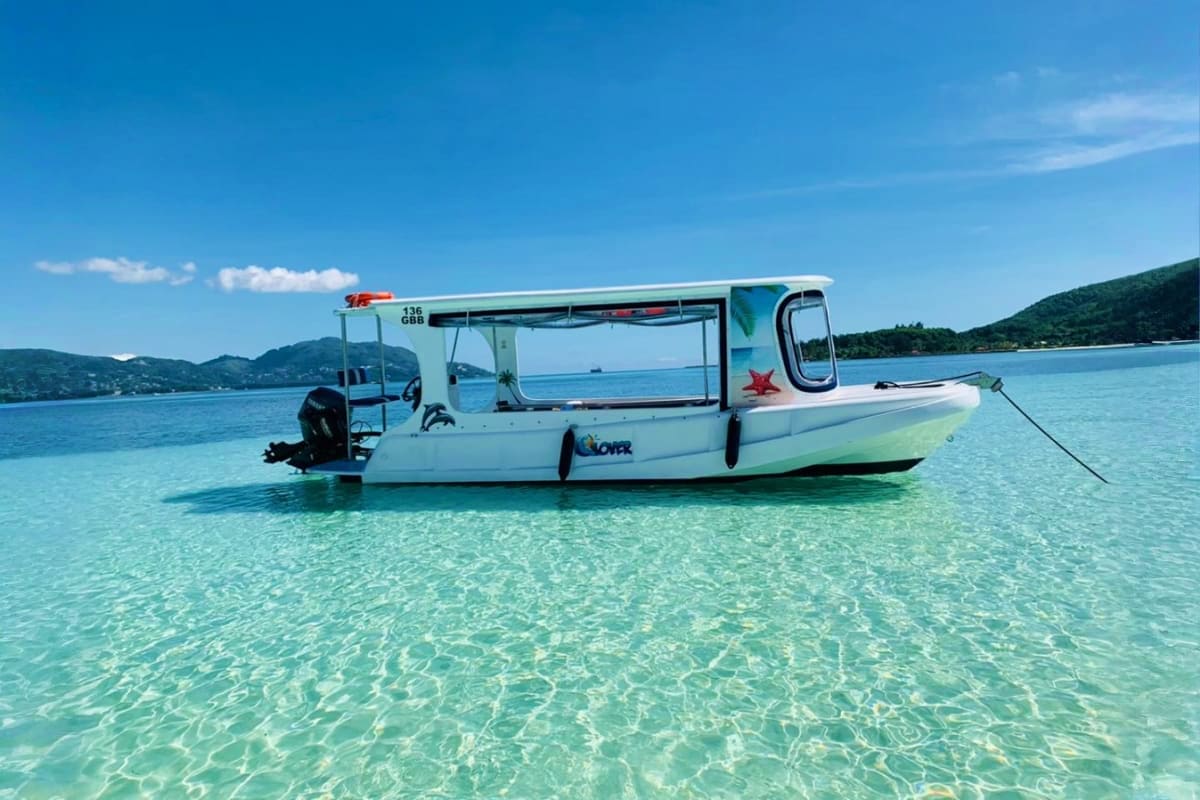 White glass-bottom boat anchored in crystal-clear turquoise water with forested mountains in the background under blue sky