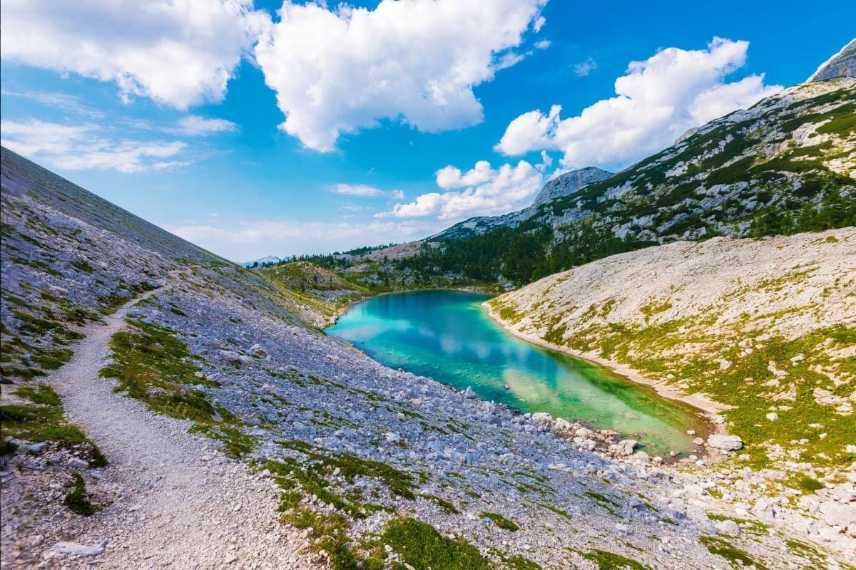 Alpine mountain lake with turquoise water surrounded by rocky limestone peaks and green vegetation under blue sky with white 
