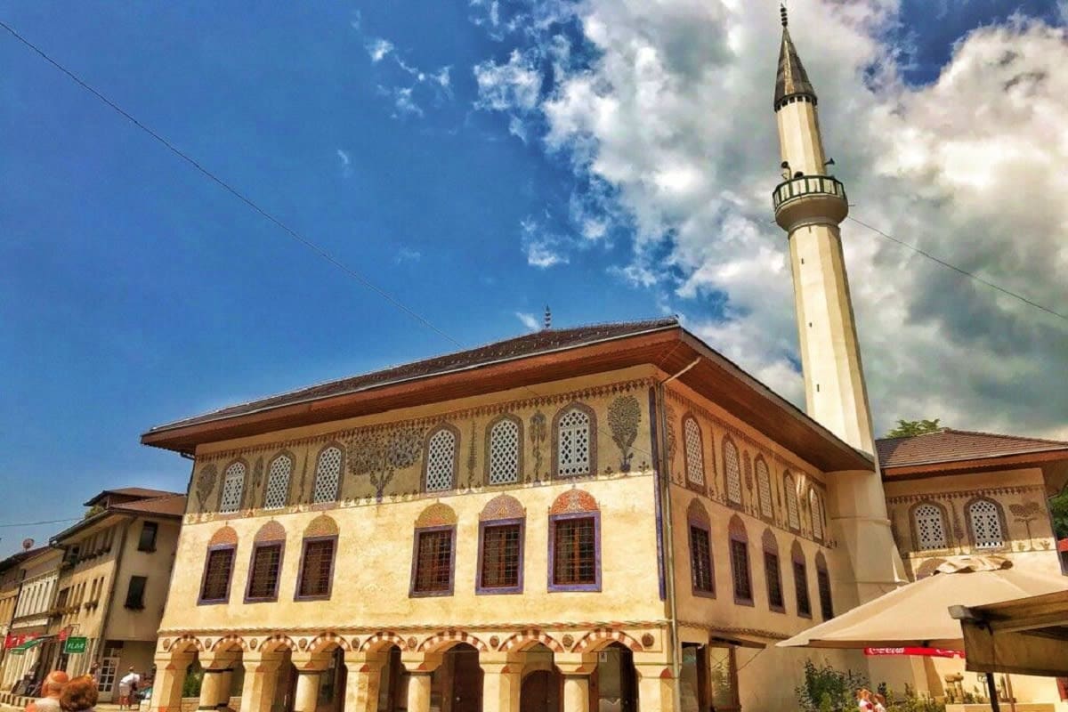 Ottoman-era mosque with cream-colored facade, decorative windows, and tall minaret against blue sky in Bosnia and Herzegovina