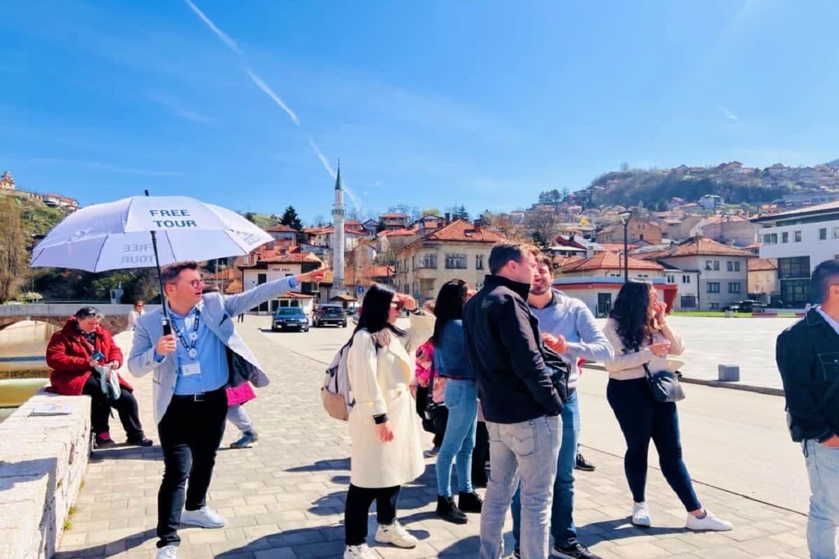 Tour guide holding free tour umbrella leading group of tourists in historic town square with mosque minaret and hillside hous