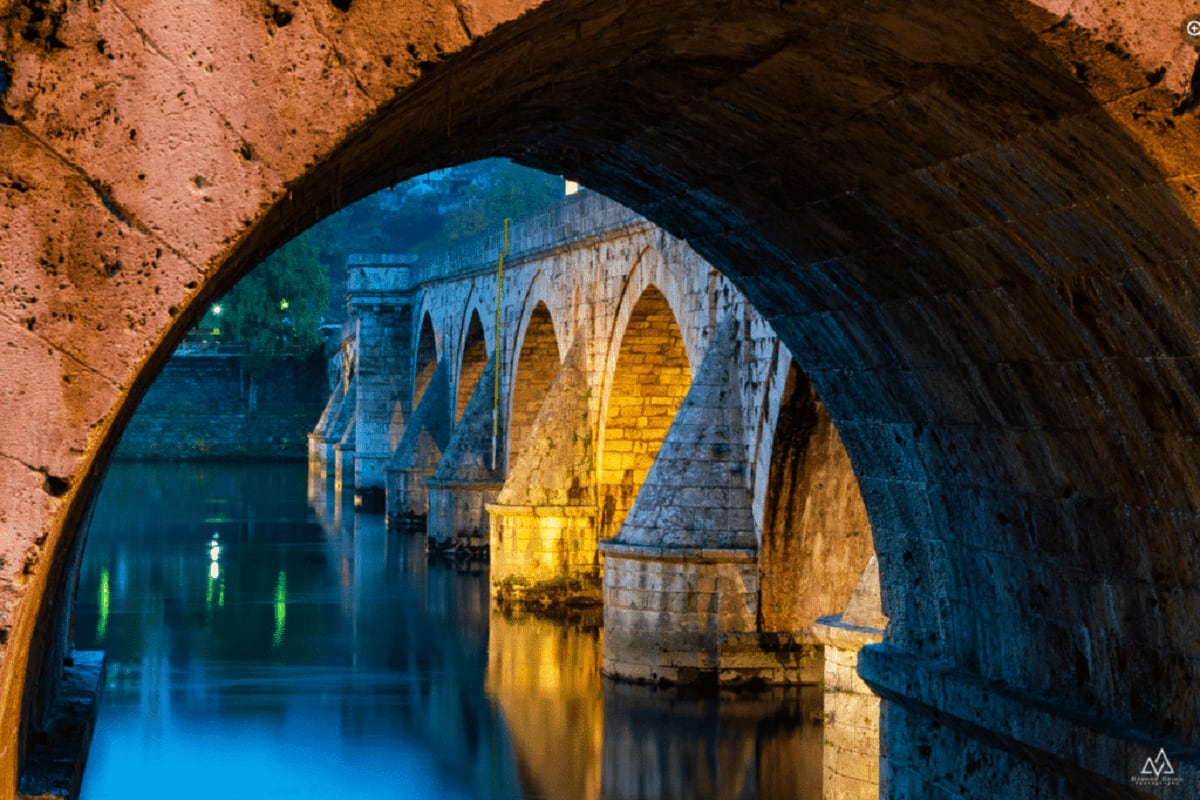 View through stone arch tunnel of illuminated historic bridge with Gothic arches spanning calm water at dusk