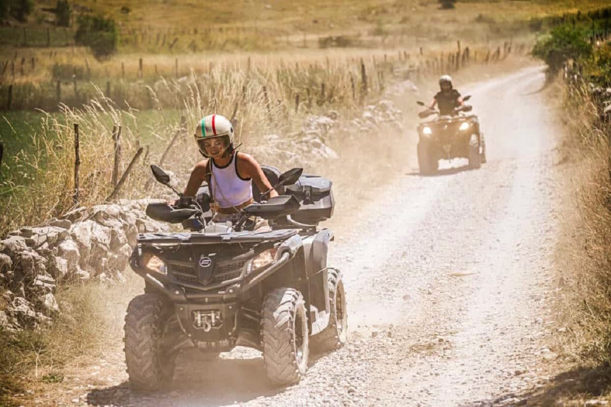 Person riding ATV on dusty dirt road through rural countryside in Bosnia, with second ATV visible in background and dry field