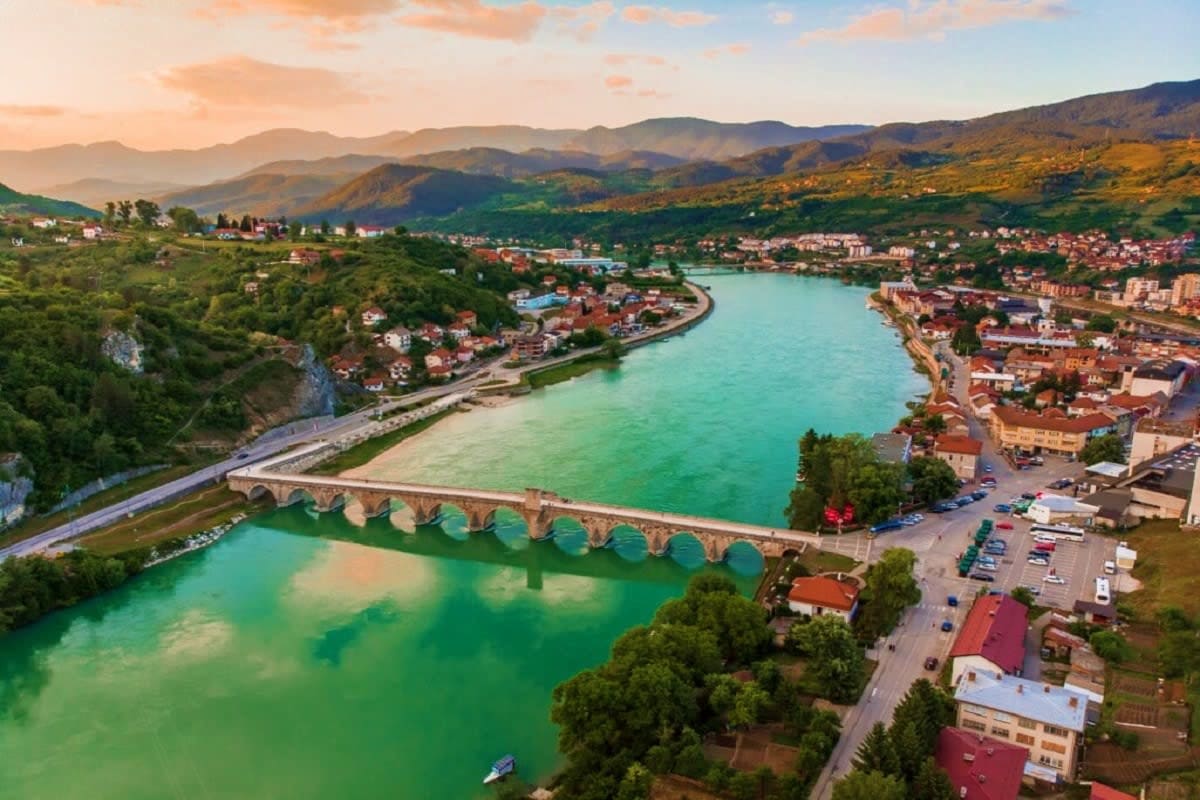 Aerial view of Konjic bridge spanning turquoise Neretva river with surrounding town, mountains, and lush green valleys at sun