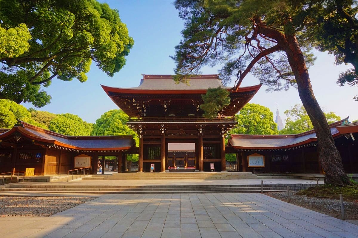 Traditional Japanese Shinto shrine with red-trimmed wooden architecture, curved roofs, stone courtyard, and tall trees under 