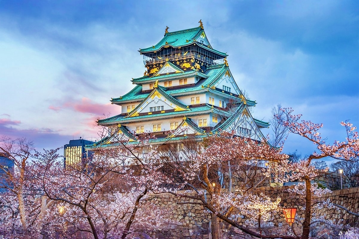 Osaka Castle with turquoise tiled roofs and gold accents surrounded by blooming cherry blossom trees at dusk