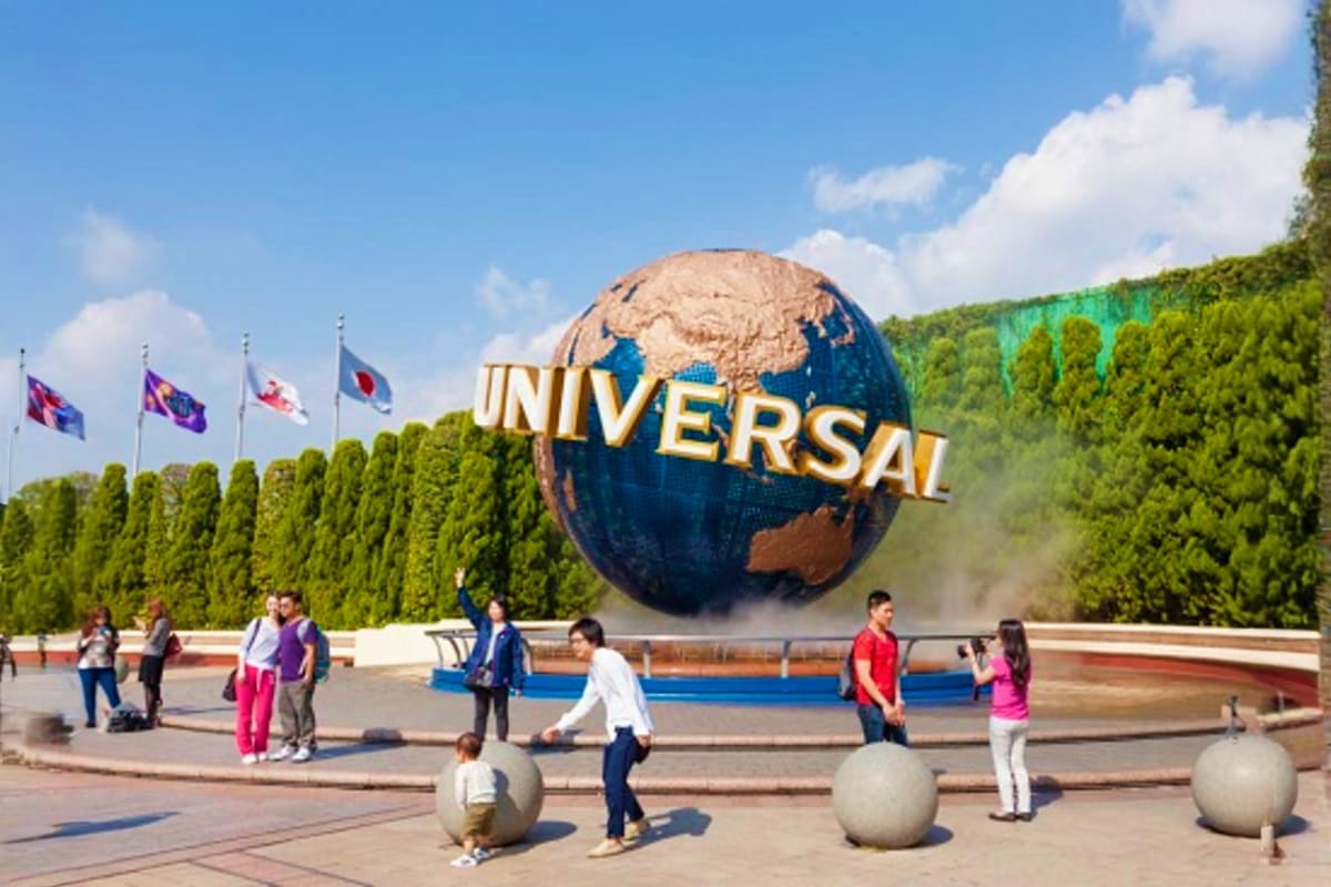 Visitors at Universal Studios Japan entrance with iconic globe logo, international flags, and forested hillside backdrop
