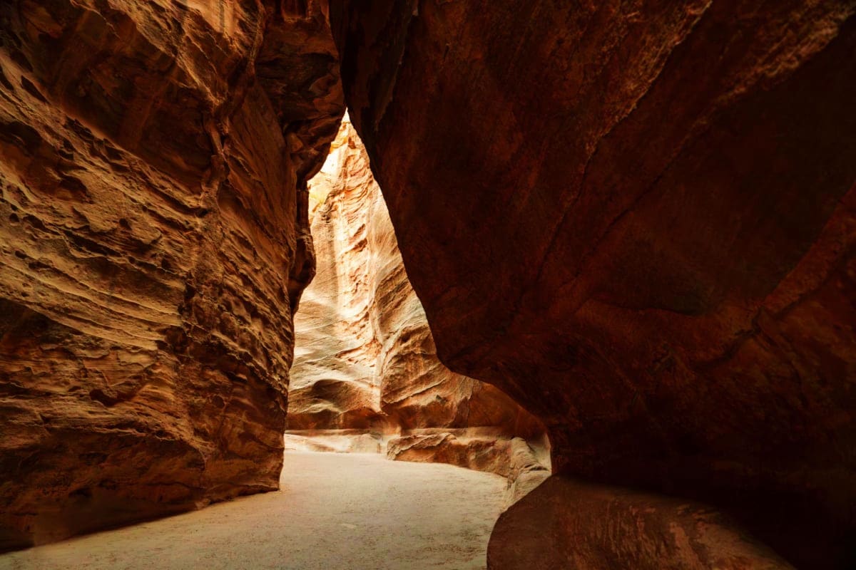 Narrow sandstone canyon with towering rust-colored rock walls and sandy floor opening to bright light ahead