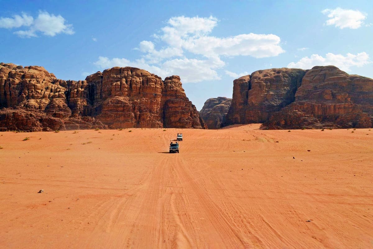Vehicles driving on red sand desert road toward towering rock formations in Wadi Rum, Jordan under blue sky with white clouds