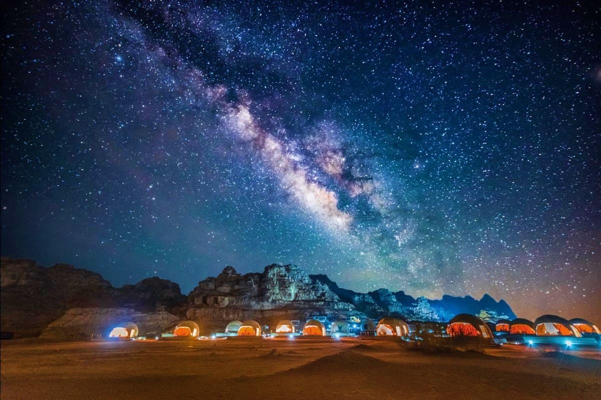 Illuminated glamping tents in desert camp beneath Milky Way galaxy and rocky mountains in Wadi Rum, Jordan at night