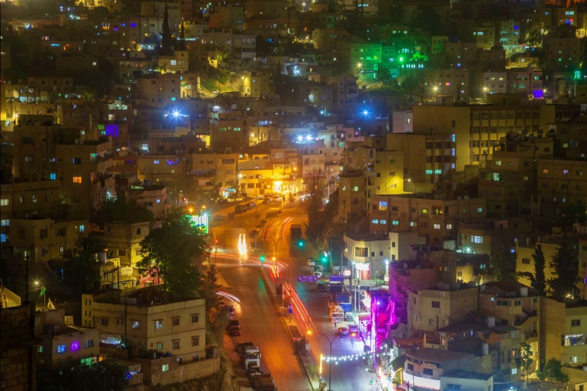 Aerial nighttime view of busy Amman street with illuminated buildings, colorful neon signs, and traffic lights creating light