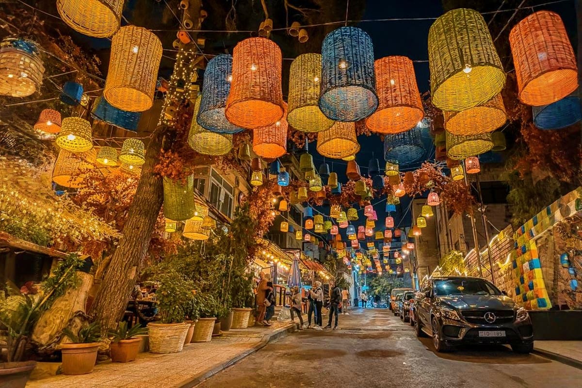 Colorful woven lanterns hanging above a narrow street at night with parked cars and pedestrians in an old town alley.