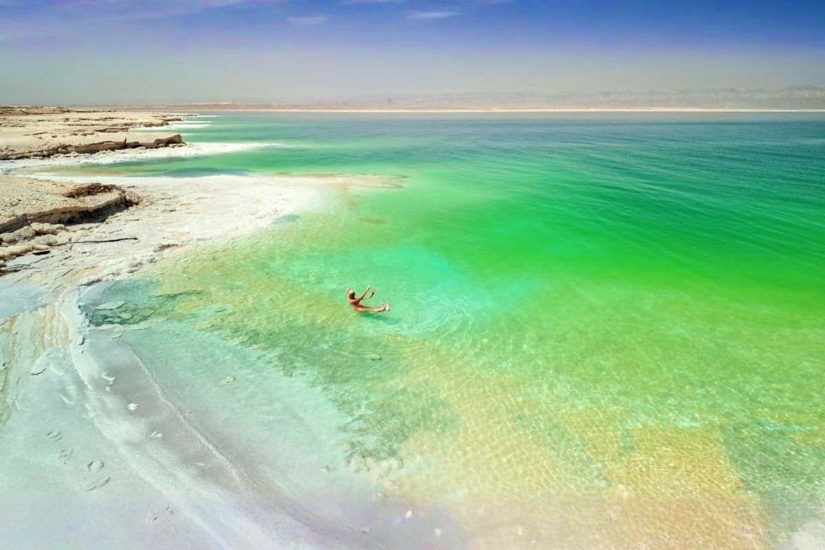 Person floating in turquoise waters of the Dead Sea with white mineral-rich shoreline and rocky cliffs in Jordan