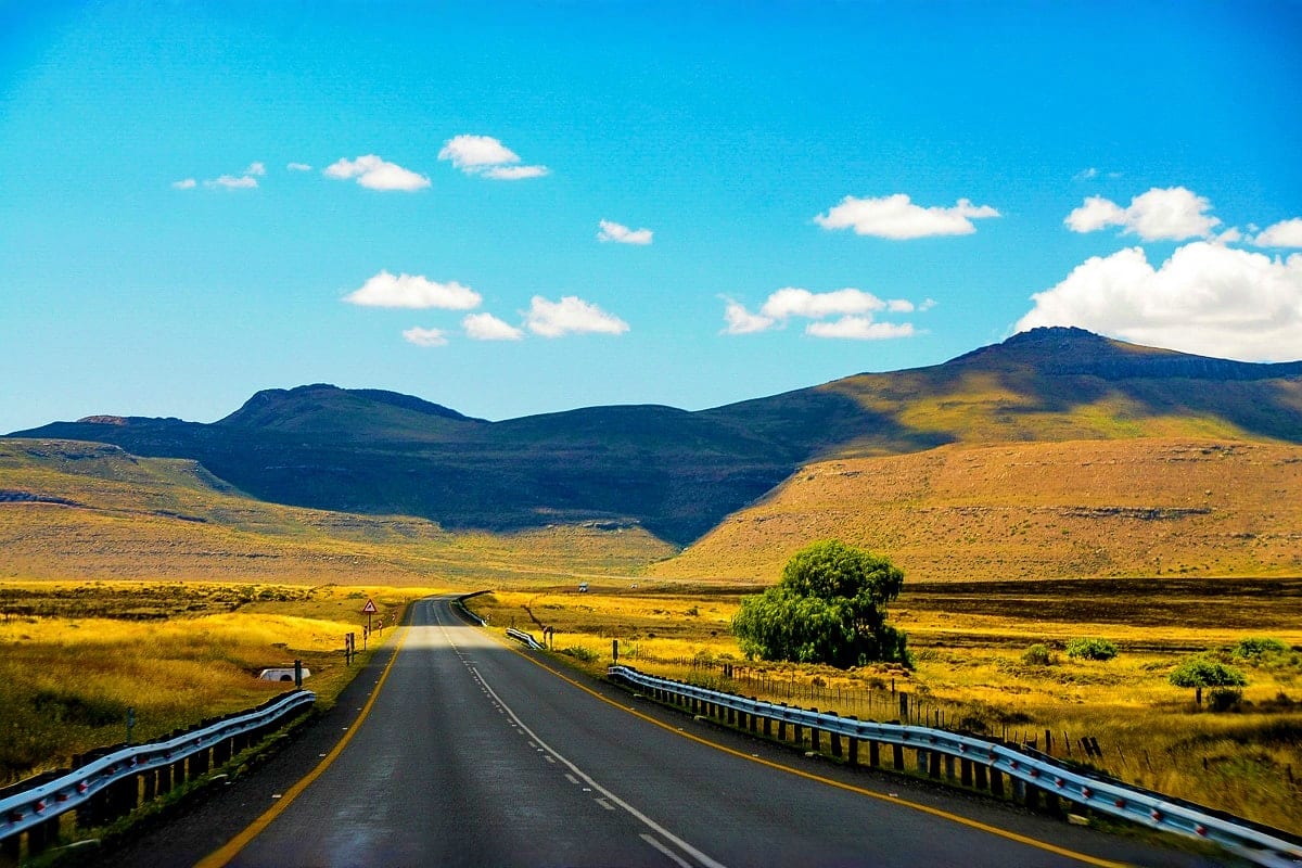 Straight road stretching through golden grassland toward volcanic mountains under blue sky with white clouds in South Africa