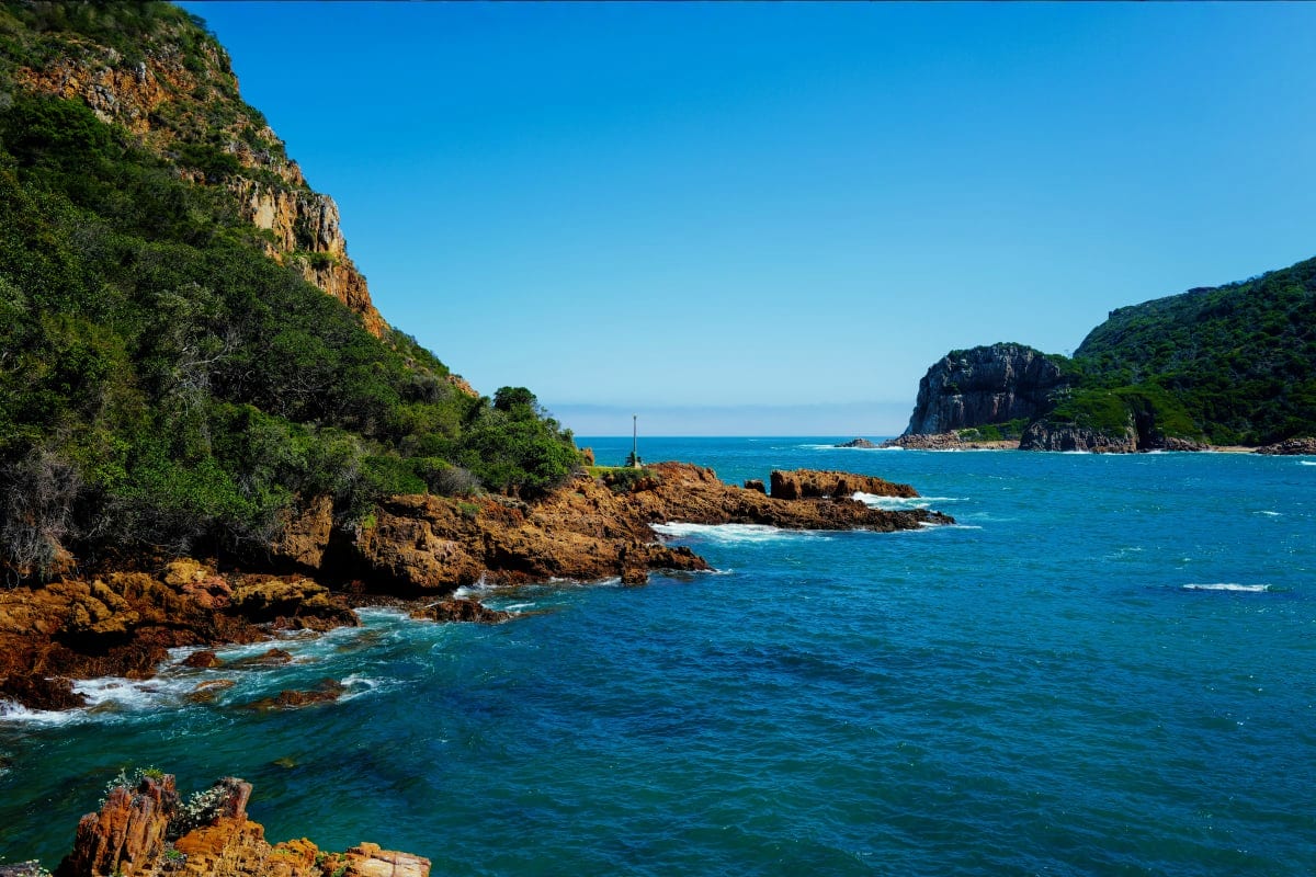 Rocky coastline with lush green cliffs, turquoise ocean waters, and distant rocky outcrops under clear blue sky