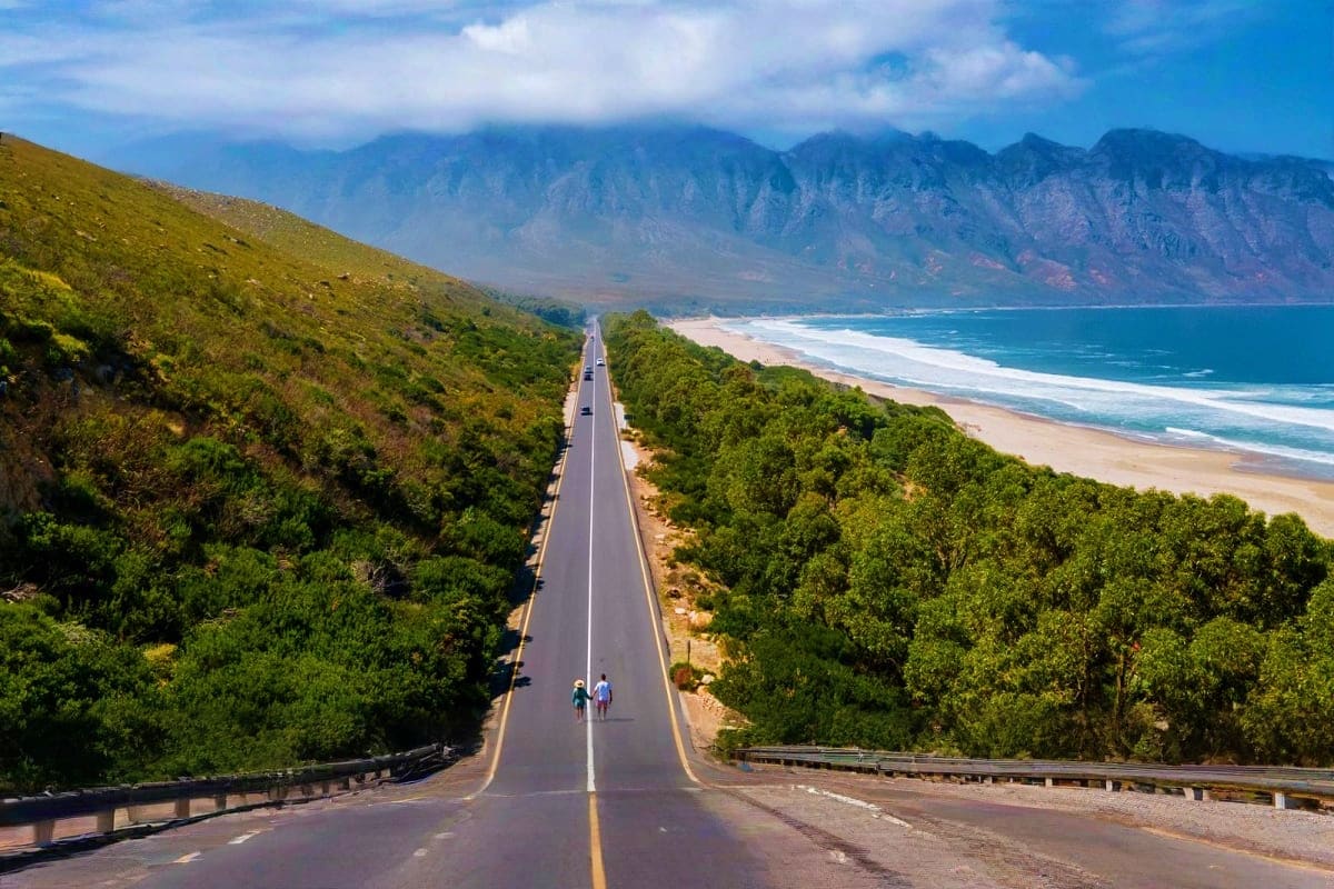 Straight road stretching toward ocean and mountains, with sandy beach and green vegetation on sides, Cape Town coastal landsc