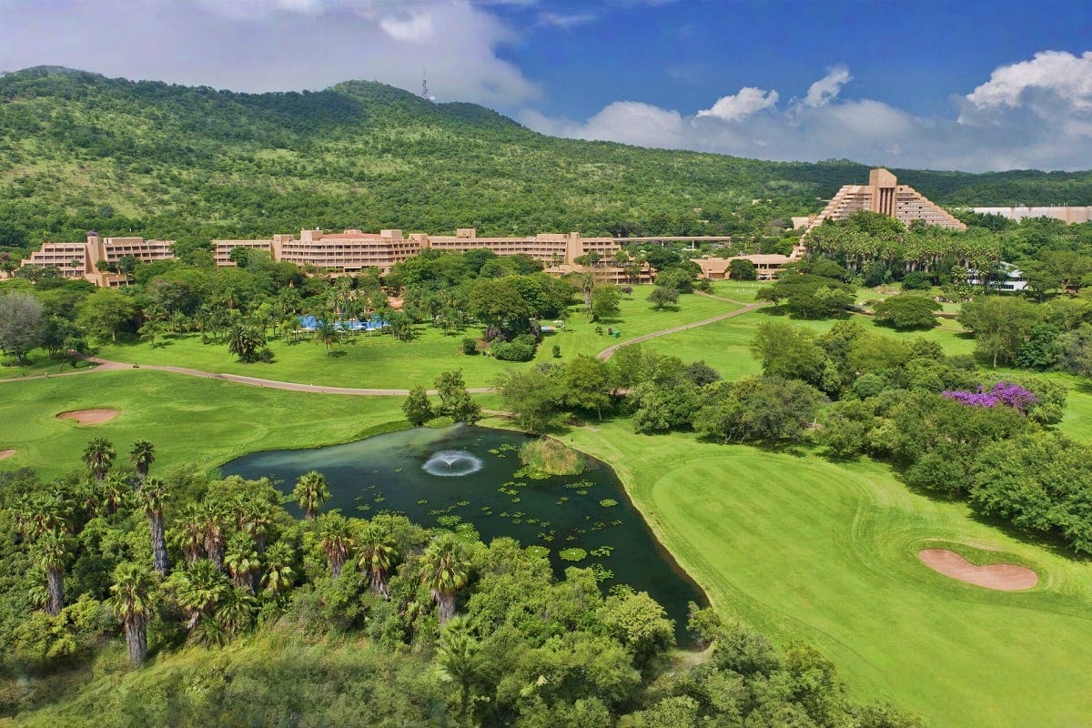 Aerial view of a large resort complex nestled in lush green hills with a golf course, palm trees, and a pond with fountain