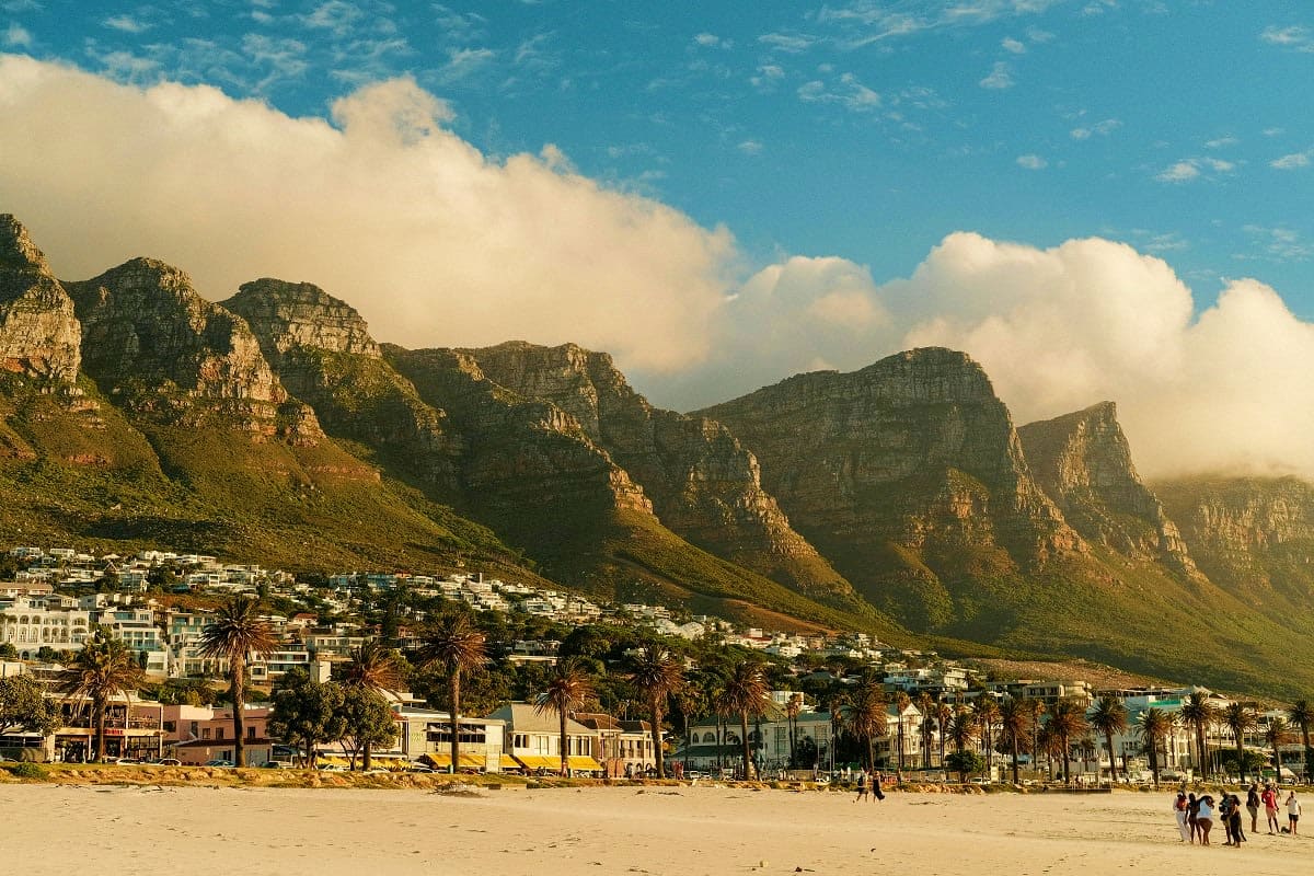 Sandy beach in Cape Town with colorful beachfront houses, palm trees, and dramatic Table Mountain peaks rising behind under b