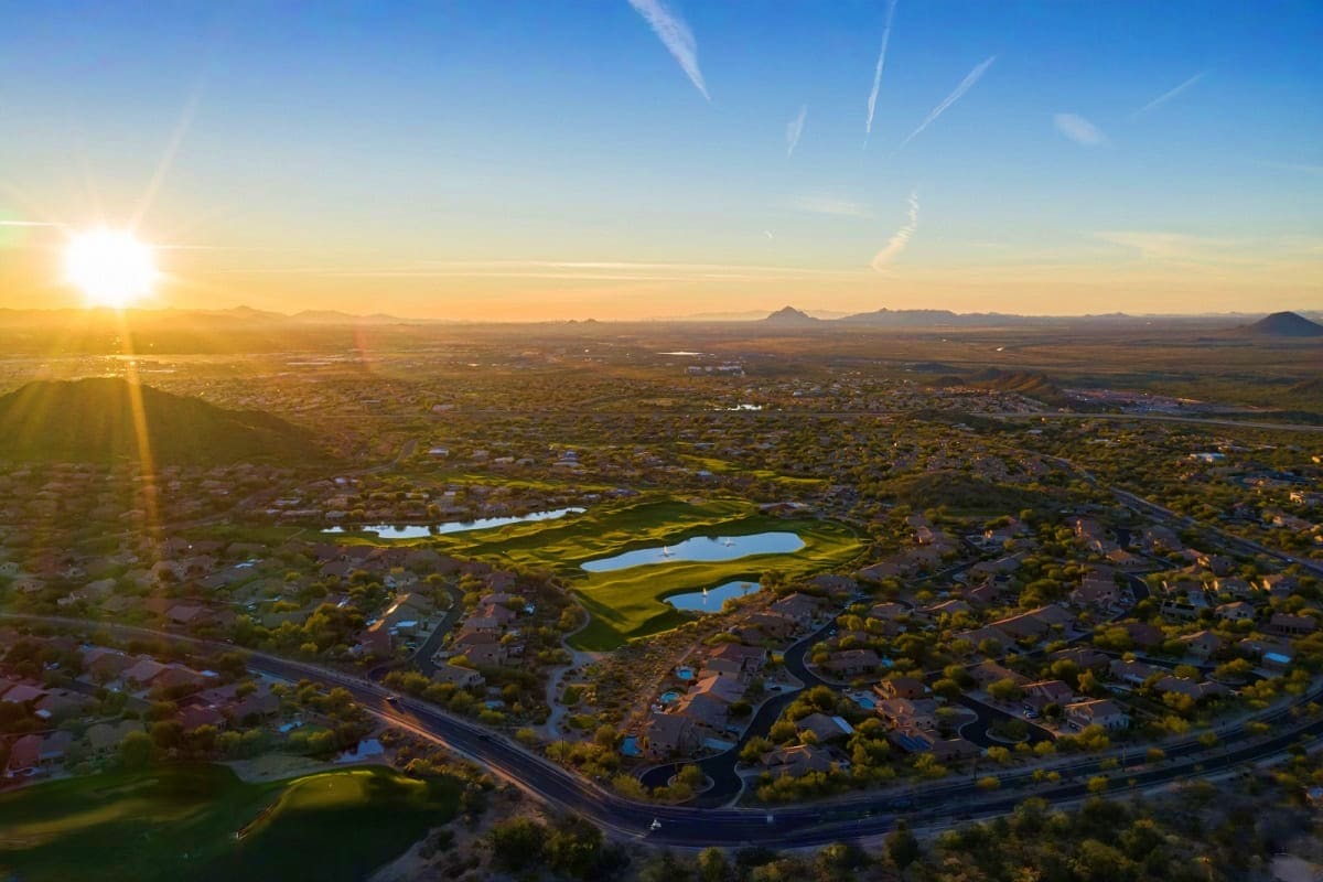 Aerial sunrise view of a golf course with blue water hazards surrounded by residential neighborhoods and distant mountains in