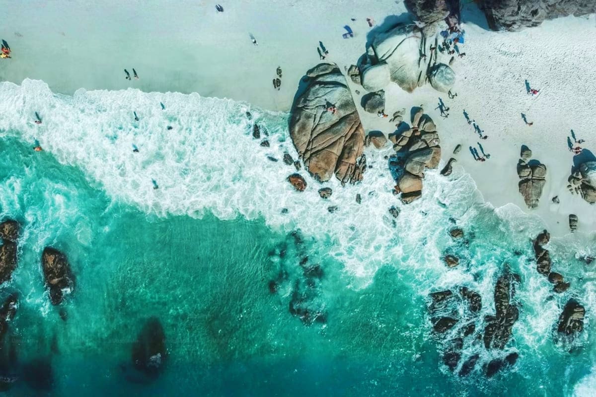 Aerial view of turquoise ocean waves crashing on white sand beach with rocky outcrops and scattered beachgoers at Cape Town c
