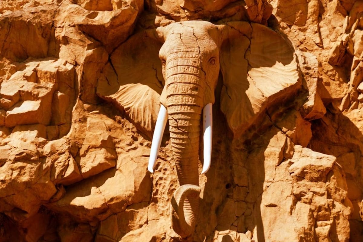 Carved wooden elephant head with white tusks mounted on terracotta rocky cliff face