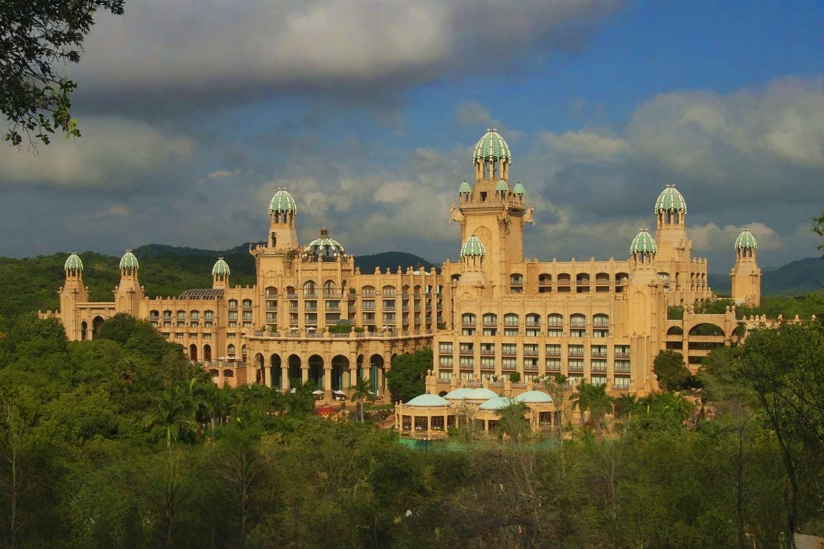 Grand ornate palace hotel with green copper domes nestled in lush mountainous landscape under cloudy sky