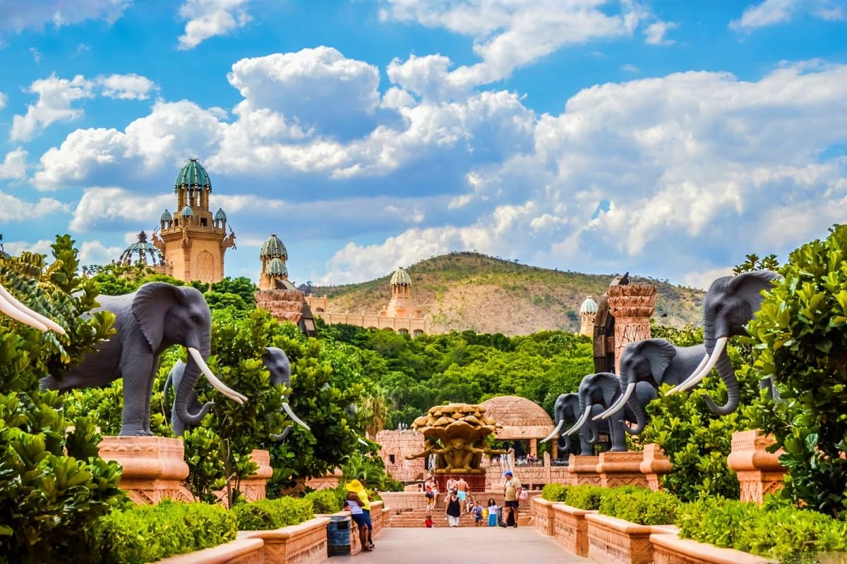 Lost City with elephant statues flanking a pathway, lush vegetation, and ancient architectural structures under cloudy blue s