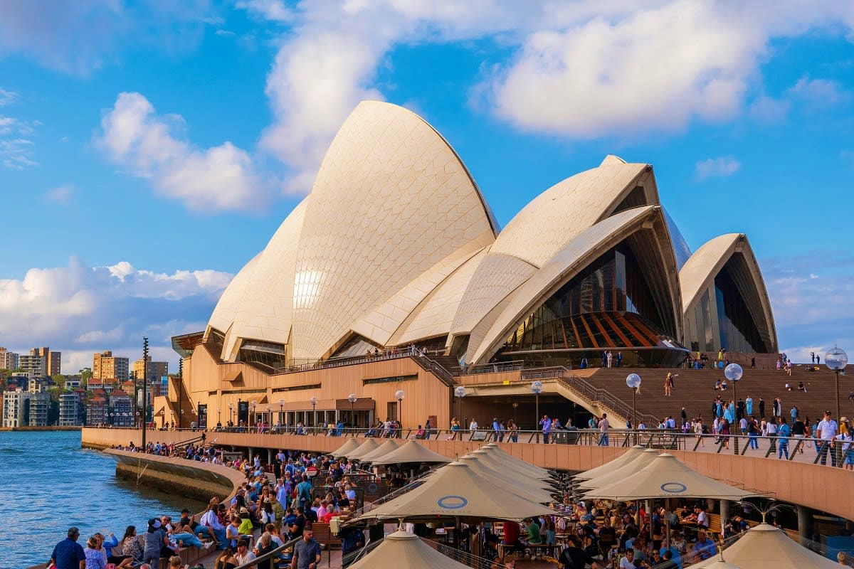 Sydney Opera House with distinctive white shell-shaped roof structures, blue harbor water, and crowds of visitors on the fore