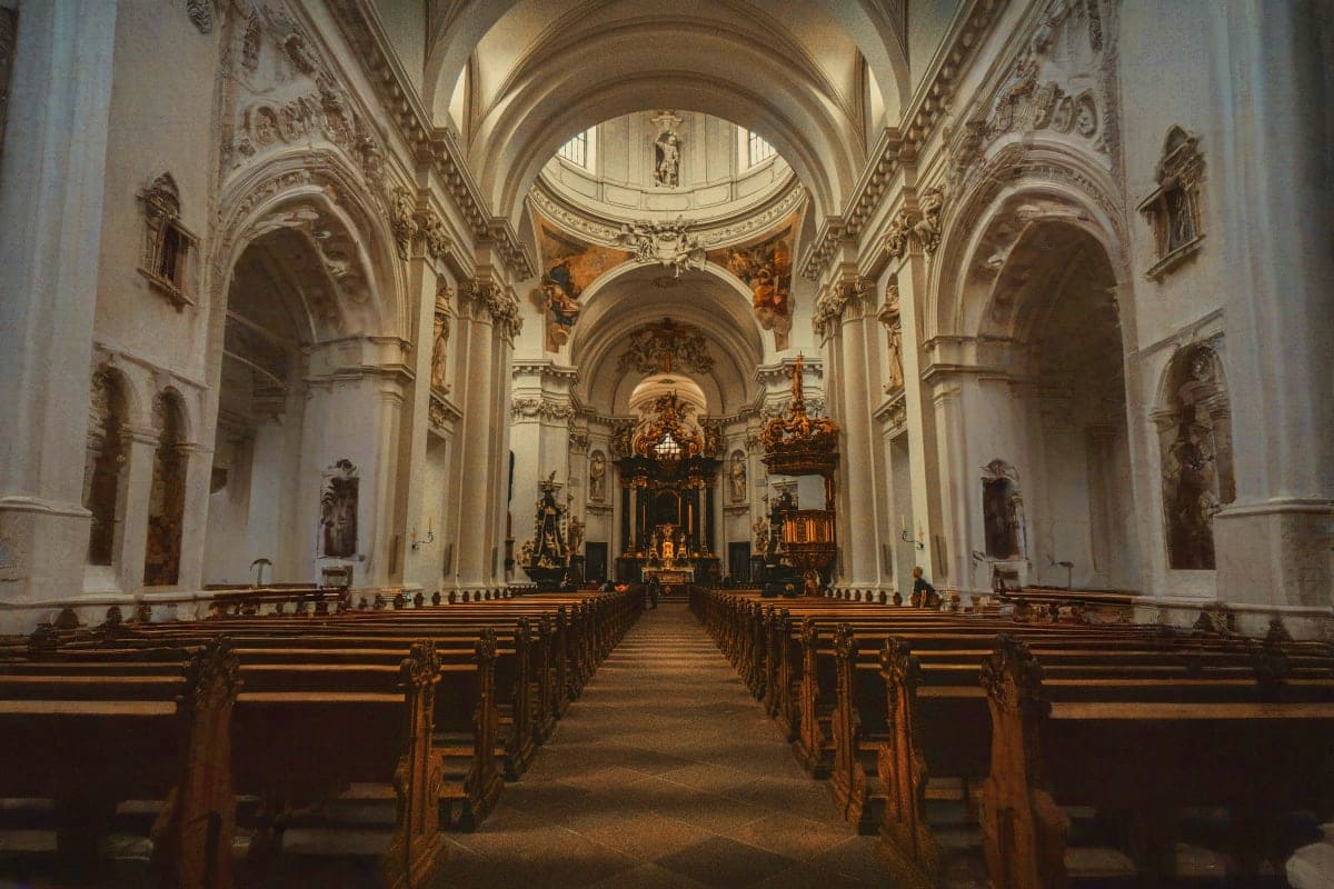 Interior of baroque cathedral with ornate white arches, wooden pews, and golden altar beneath domed ceiling