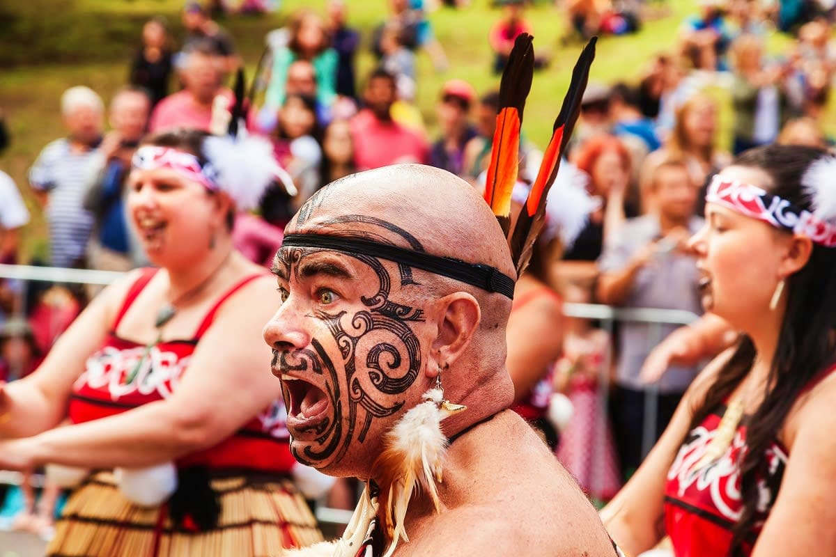 Maori man with traditional face tattoo and feathered headdress performing at cultural gathering with spectators in background