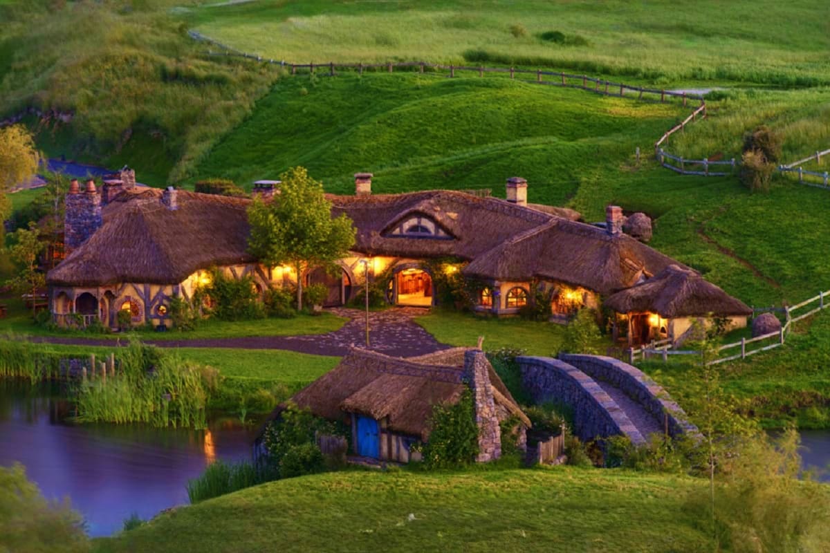 Hobbiton movie set featuring illuminated hobbit holes with thatched roofs nestled in rolling green hills beside a stream in N
