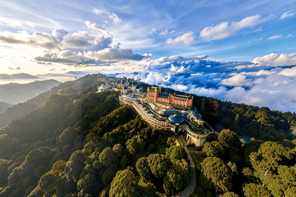 Aerial view of Pena Palace perched on a forested mountain ridge in Portugal with clouds below and blue sky above