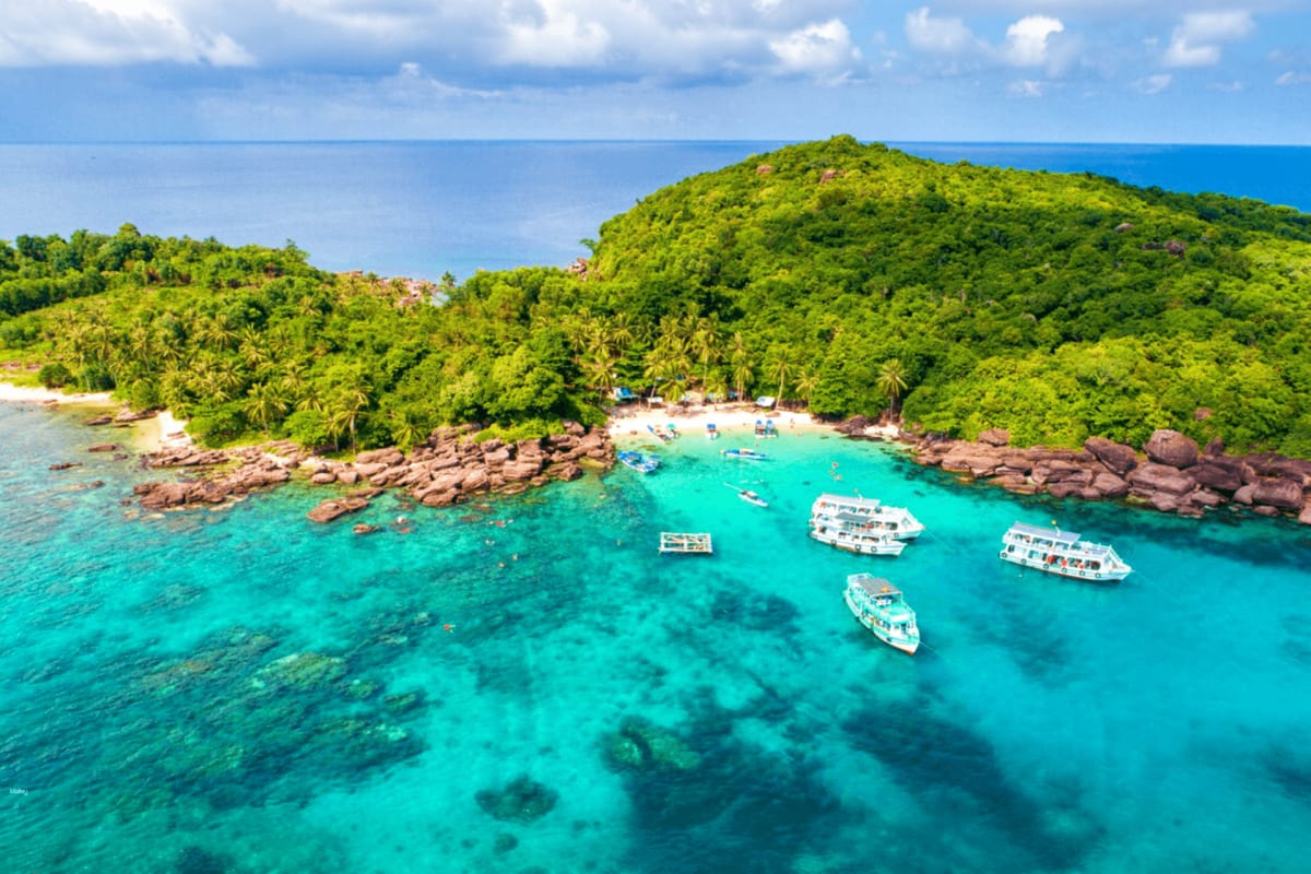 Aerial view of tropical island cove with turquoise water, moored boats, white sand beach, and lush green forested hills under