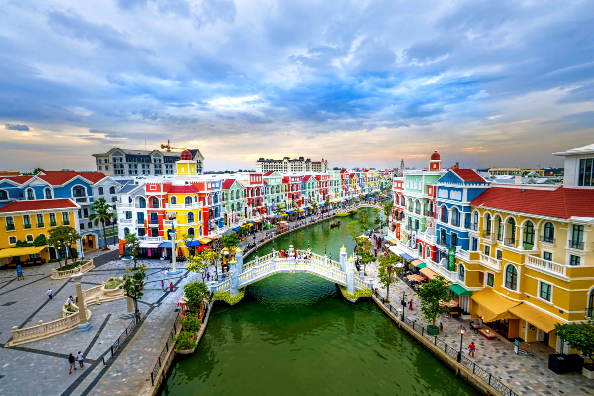 Colorful Venice-inspired waterfront town with red-roofed buildings, arched bridge, and green canal water under cloudy sky