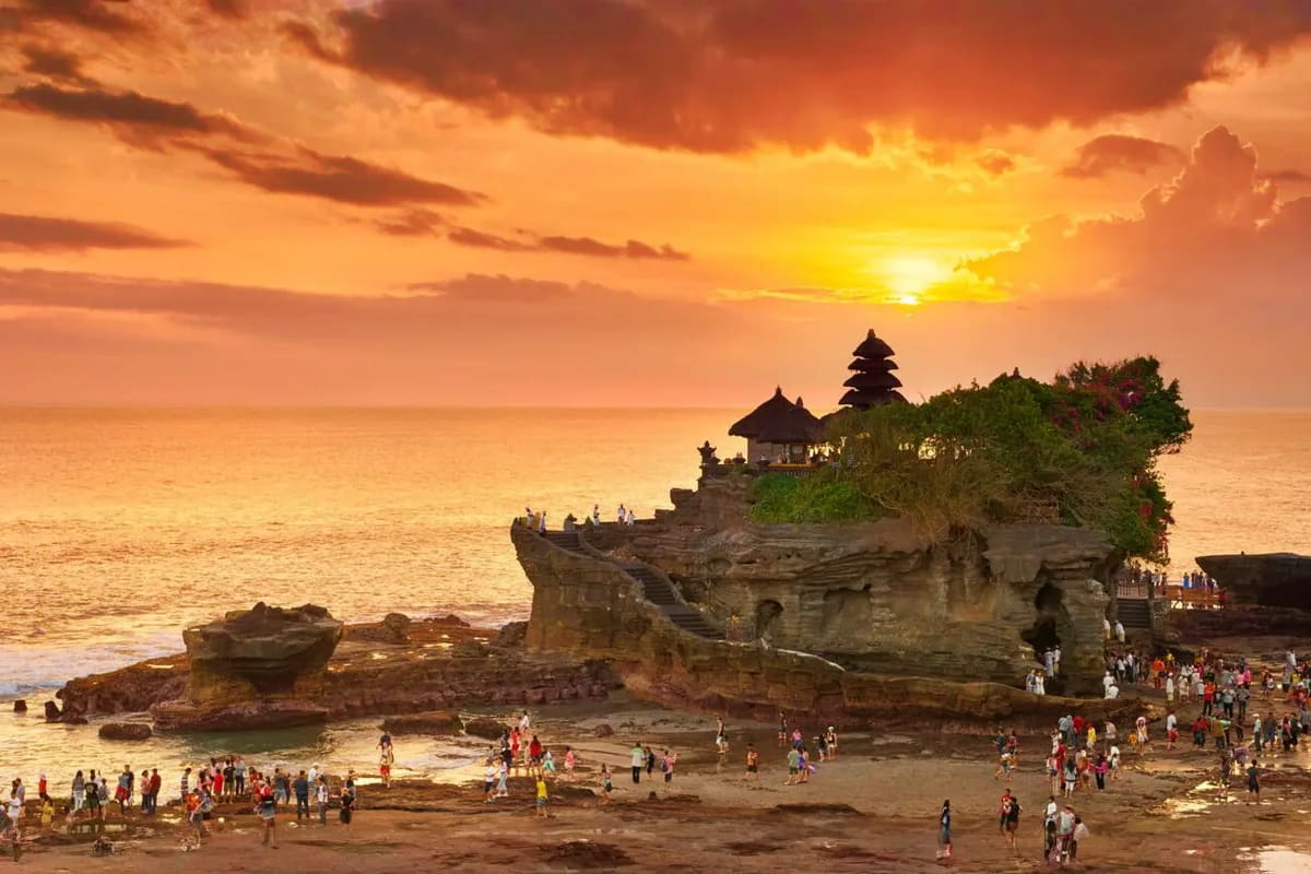 Tanah Lot temple perched on rocky coastal cliff at sunset with tourists on beach and golden sky in Bali