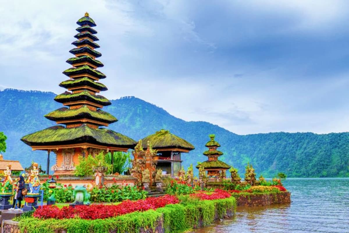 Traditional Balinese temple with moss-covered tiered roofs on a lake shore, surrounded by red flowers and lush green mountain