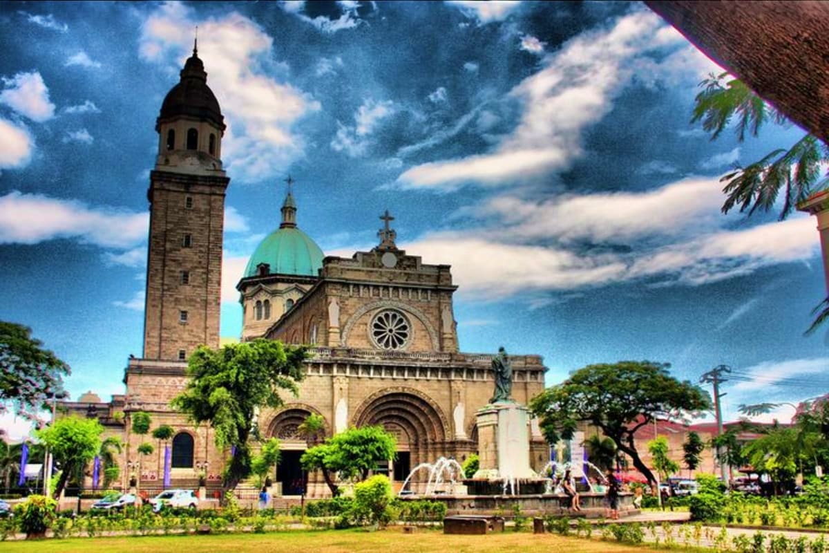 Best of Manila and Boracay Holiday Package Historic stone cathedral with bell tower and green dome under dramatic blue sky, surrounded by tropical gardens and fountain