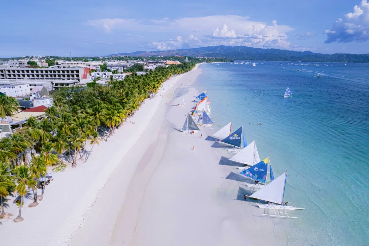 Tropical Philippines Vacation Aerial view of white sand beach with traditional outrigger boats lined up on shore, palm trees, resort buildings, and blue oc