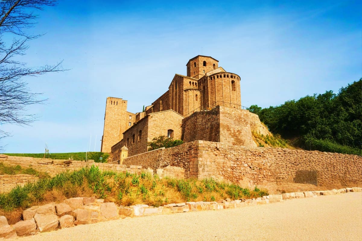 Ancient brick church with domes and towers on hilltop surrounded by stone walls and green vegetation under clear blue sky