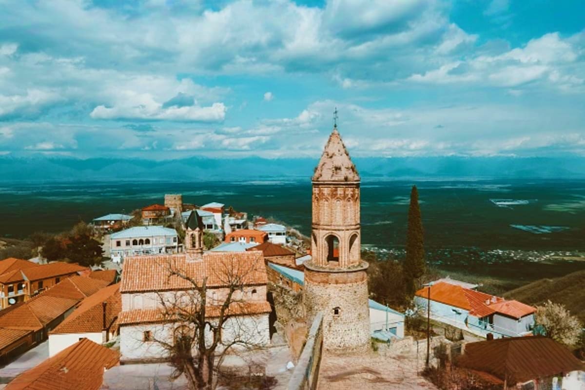 Medieval stone tower and white buildings with orange roofs overlooking vast green valley and mountains under cloudy blue sky