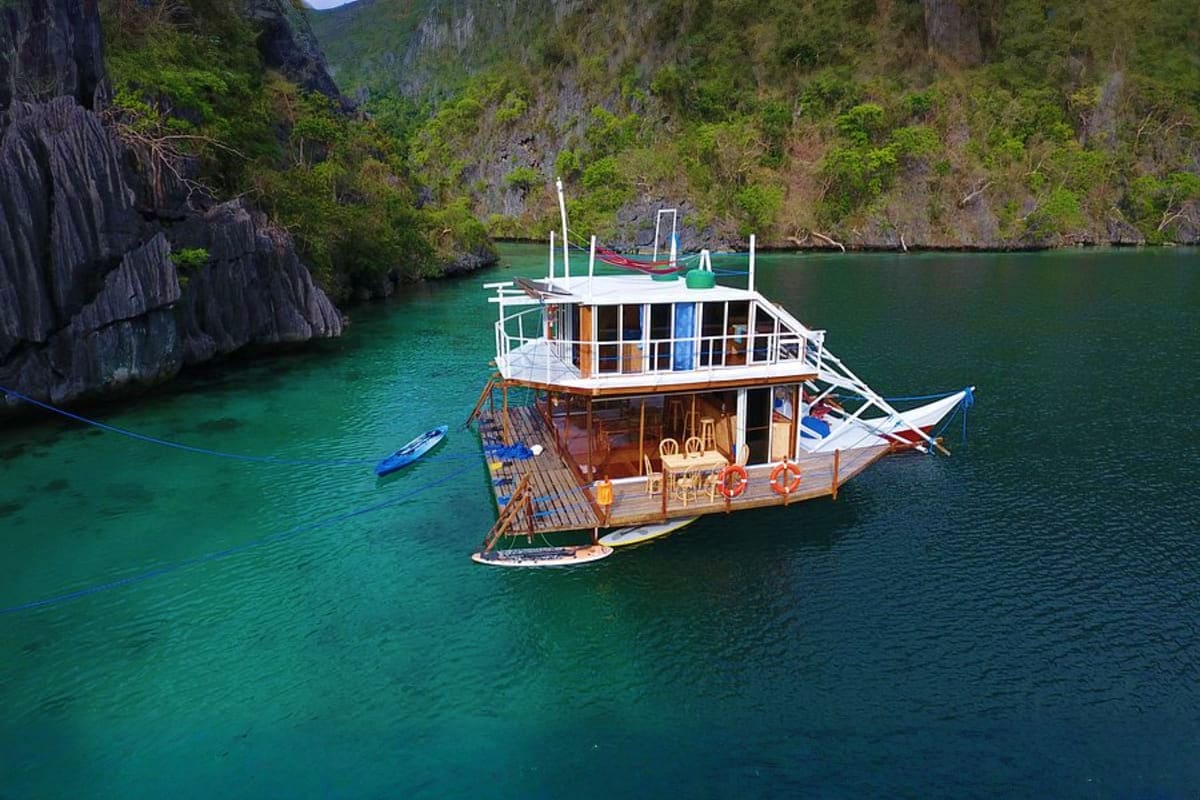 Beyond Manila Traditional wooden tour boat anchored in turquoise lagoon surrounded by tall limestone cliffs and lush vegetation