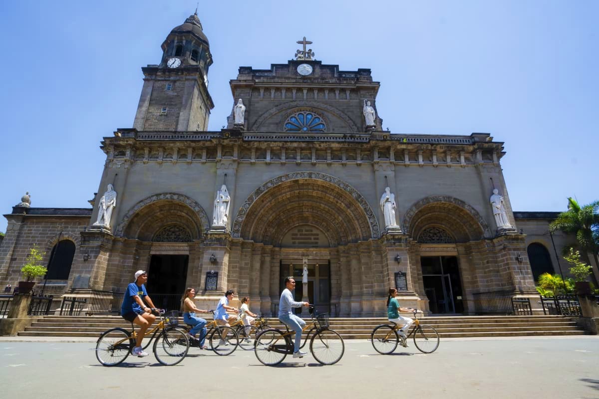 Beyond Manila Group of cyclists riding past historic stone cathedral with clock tower and ornate arched entrances in tropical setting