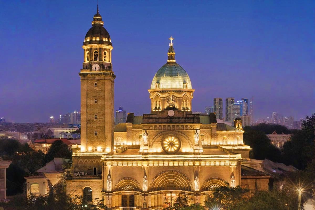 Illuminated basilica with golden dome and bell tower at dusk, Manila cityscape in background