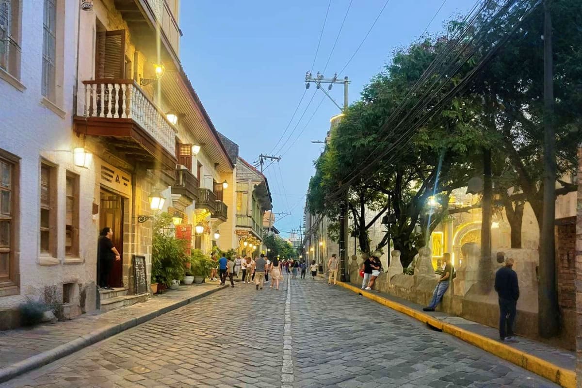 Historic cobblestone street in Manila's Intramuros district at dusk with colonial buildings, street lights, and pedestrians w